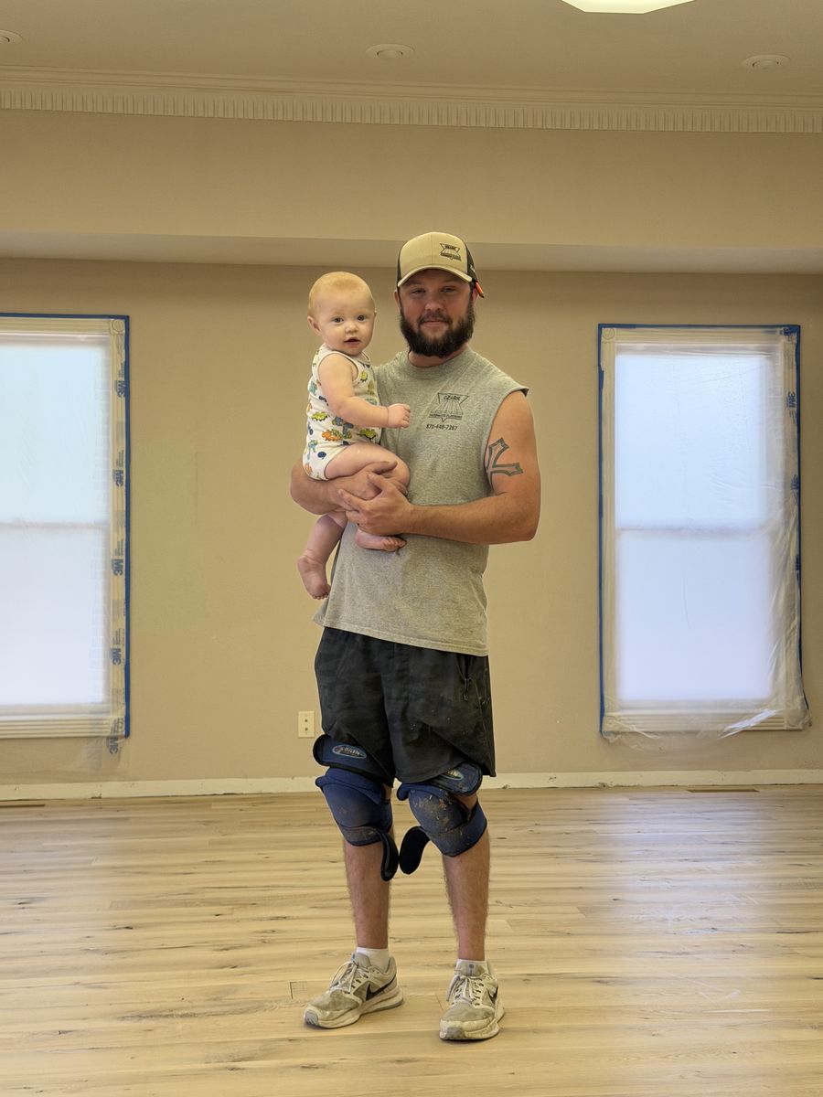 Ethan Walsh in an Ozark Hardwood Flooring shirt and knee pads holding his baby son Winston on a newly finished floor